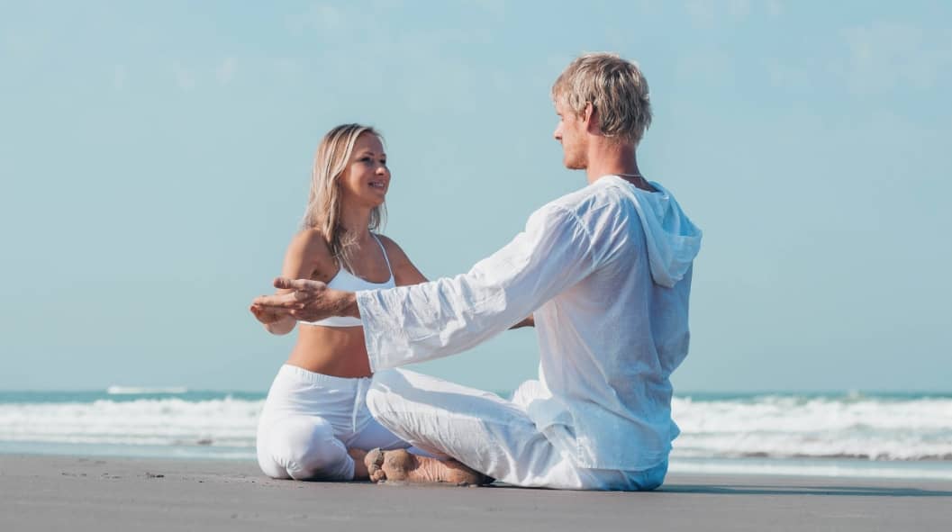 guy and girl in one of sober date ideas wearing white clothes are sitting on the ocean and holding hands doing yoga
