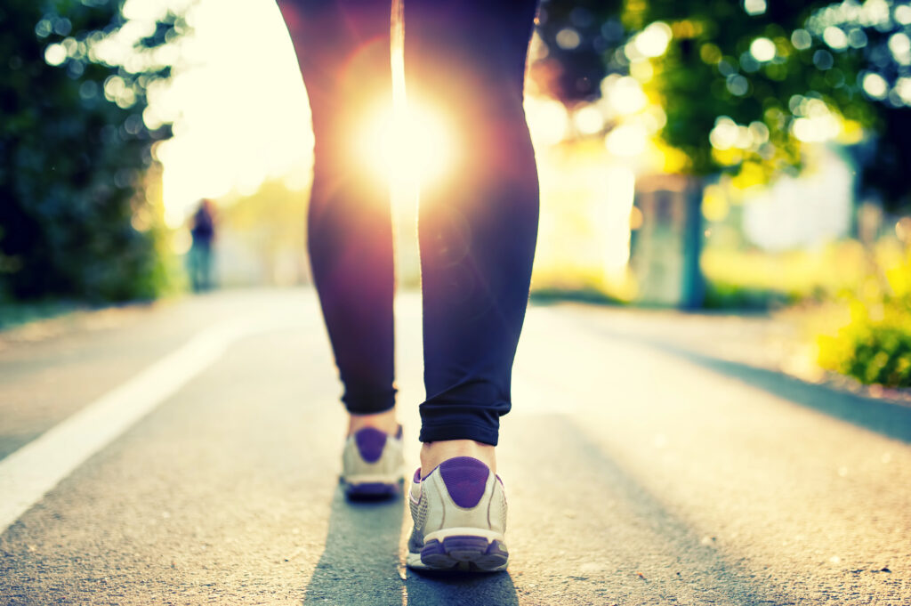 Person walking on a paved path in athletic shoes at sunrise or sunset, with sunlight shining between their legs and trees in the background.