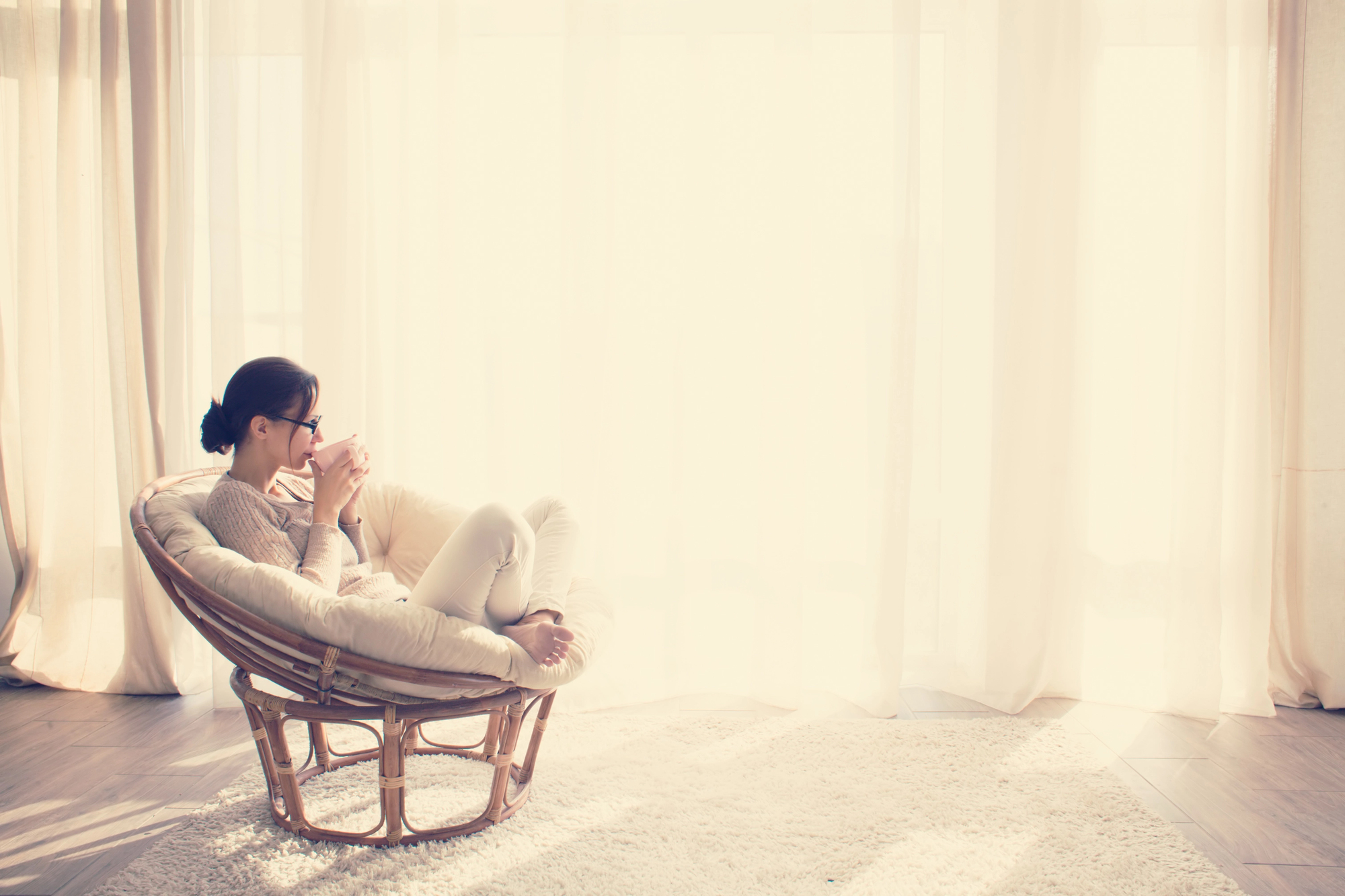 A person sits in a round wicker chair, holding a mug, by a large window with sheer curtains in a softly lit room.