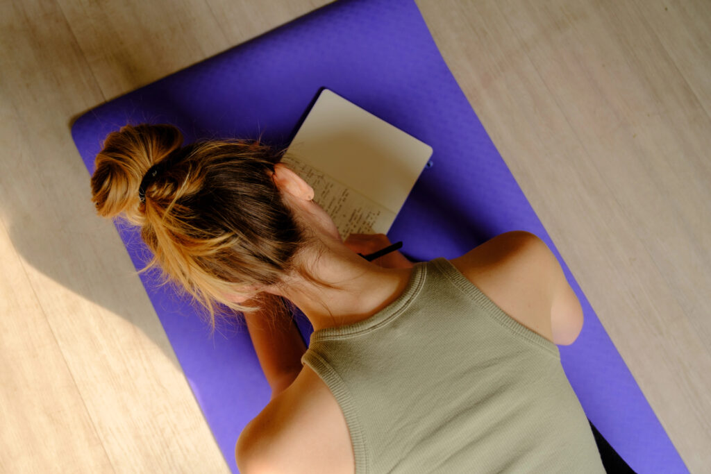 A person with light brown hair in a bun writes in a notebook while sitting on a purple yoga mat, viewed from above.