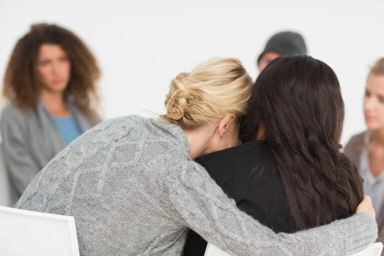 A woman with blonde hair hugs another woman in a support group setting, while three other people sit in the background.