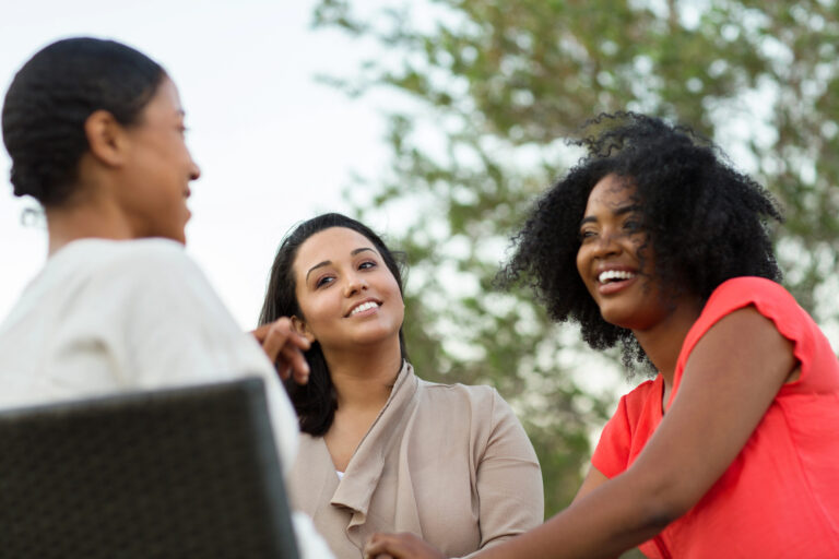 Three women sit outdoors, talking and smiling with trees in the background.