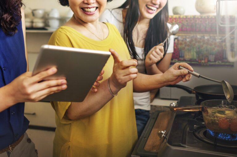 Three women in a kitchen cooking together, one holding a tablet, while another stirs a pot on the stove, all appearing engaged and cheerful.