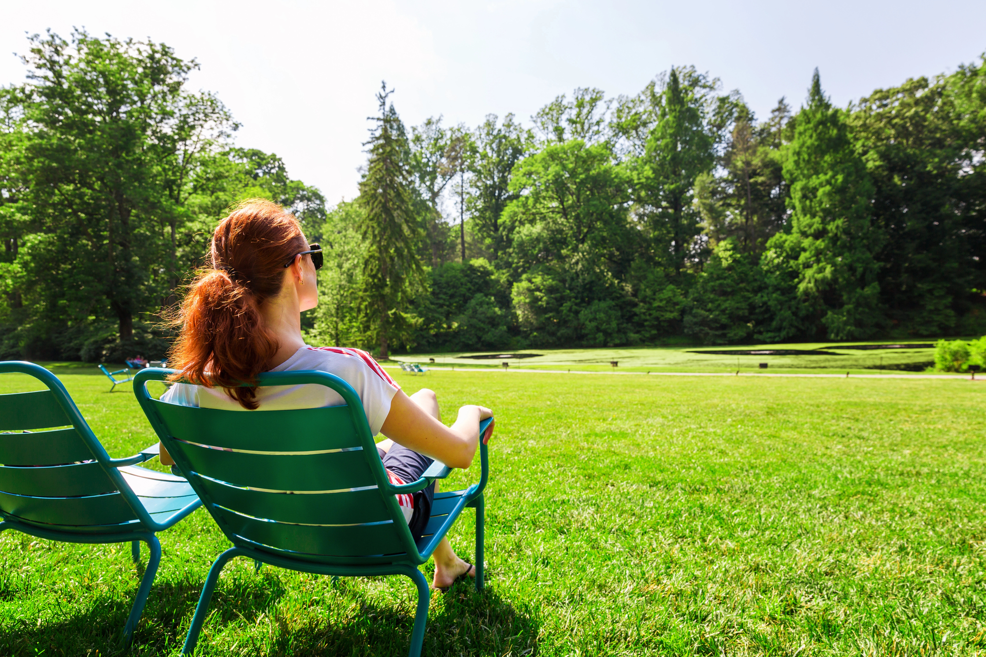 A person with brown hair sits on a metal chair in a grassy park, facing trees, on a sunny day.
