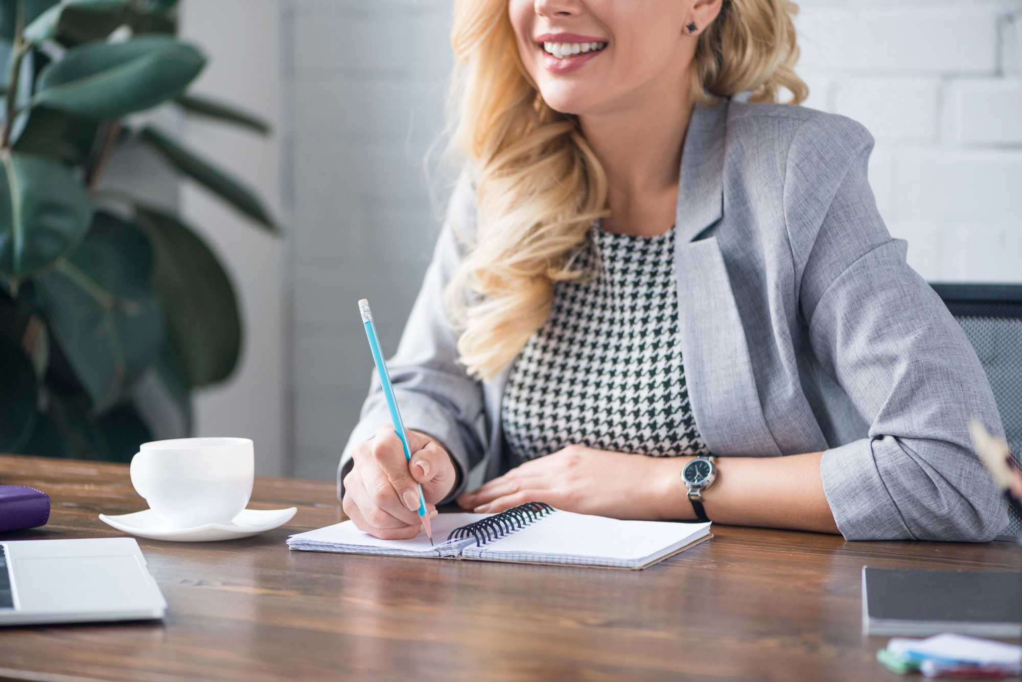 Woman in business attire sits at a desk, smiling and writing in a notebook, with a coffee cup and a potted plant nearby.