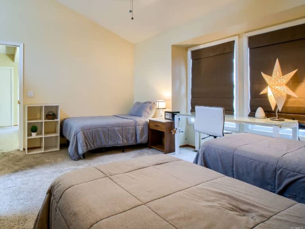 A bedroom with two twin beds, a desk with a chair, a lamp shaped like a star, two windows with dark blinds, and a small shelving unit.