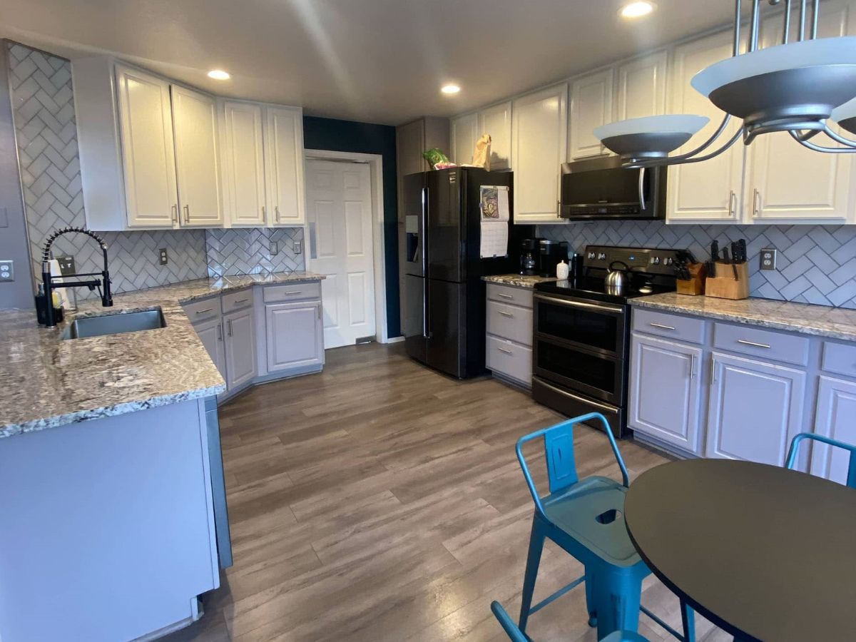 Modern kitchen with white cabinets, black appliances, gray countertops, and wood-look flooring; round dining table with blue metal chairs in the foreground.