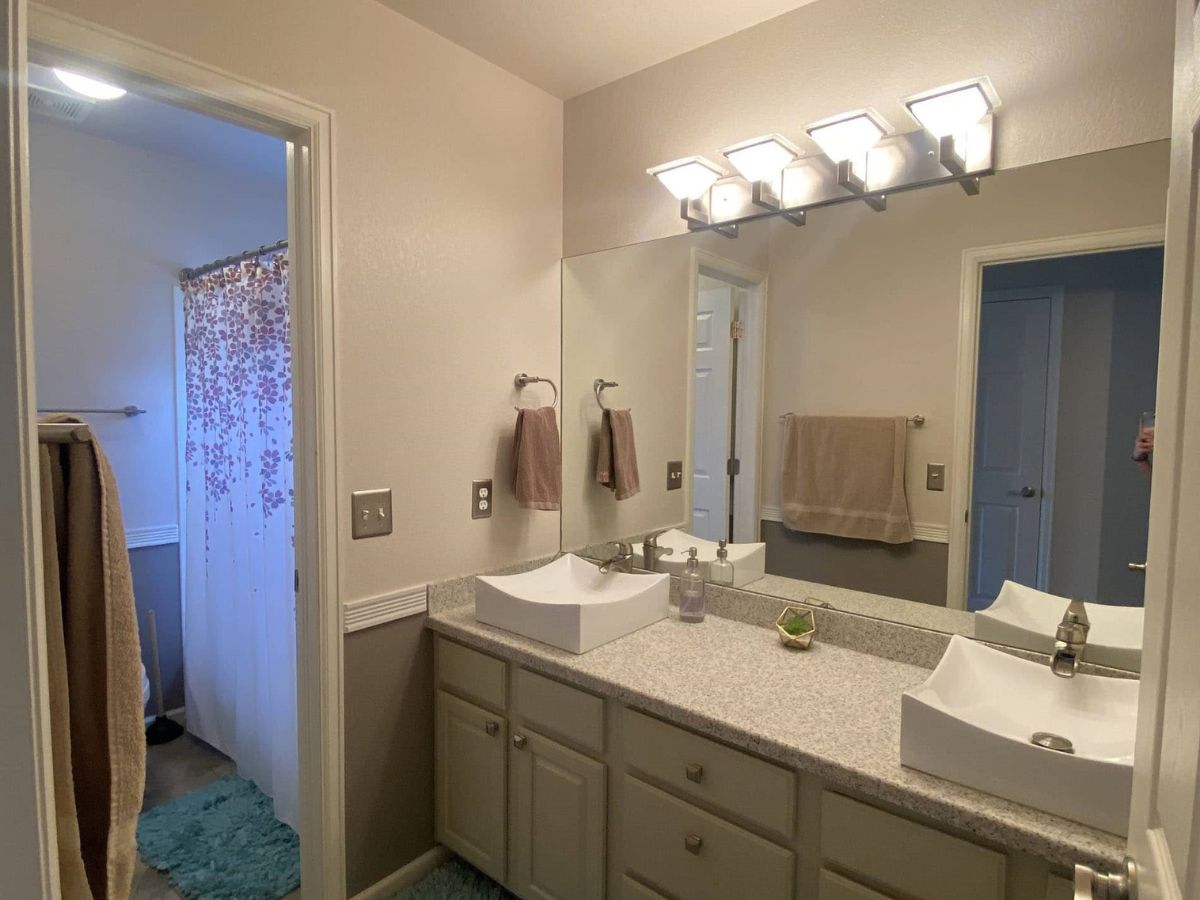 A bathroom with a double sink vanity, a large mirror, modern light fixture, tan walls, and a view into a shower area with a floral curtain and teal bath mat.