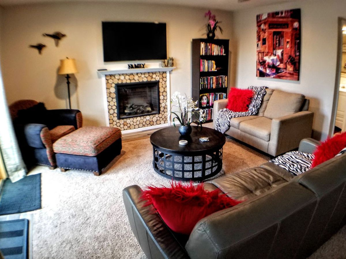 A cozy living room with a fireplace, wall-mounted TV, bookshelves, armchair, ottoman, beige sofa, gray couch, round coffee table, and red accent pillows.