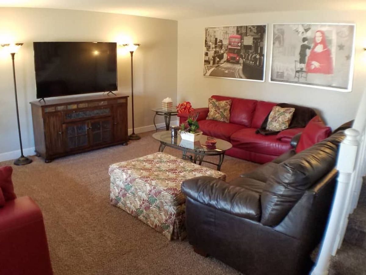 A living room with a TV on a wooden stand, red and black sofas, a floral ottoman, glass coffee table, floor lamps, and framed wall art.
