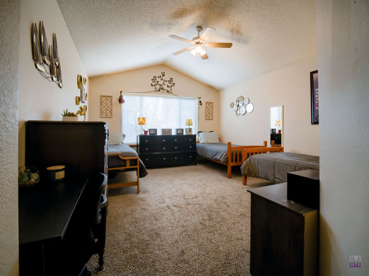 A spacious bedroom with two twin beds, two dressers, a desk, and a ceiling fan. The room has neutral walls, carpeted flooring, and natural light from a large window.