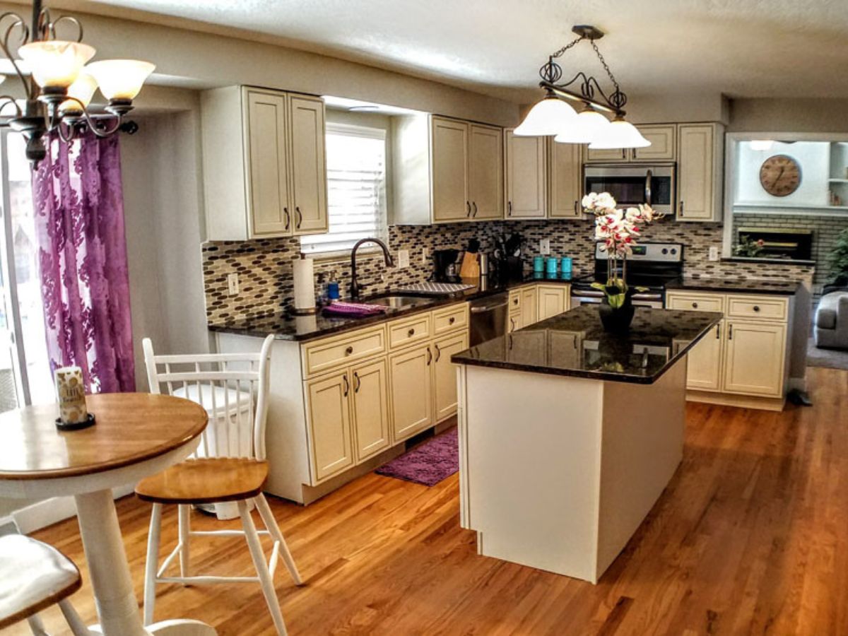 Modern kitchen with cream cabinets, black countertops, tile backsplash, island with a flower vase, round table with chairs, and hardwood floors. Purple curtains and a rug add color.