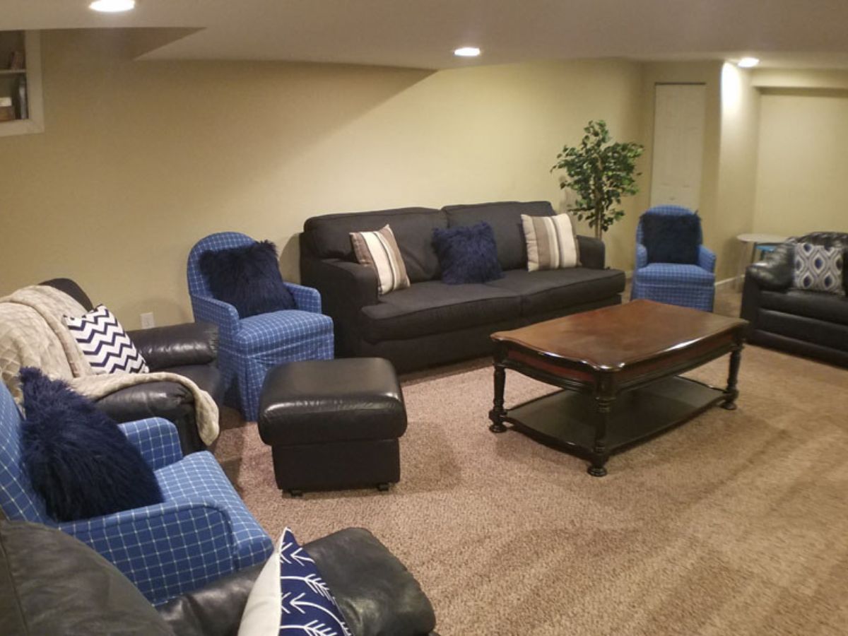 A furnished living room with a black sofa, blue armchairs, an ottoman, a wooden coffee table, and decorative pillows on a carpeted floor.