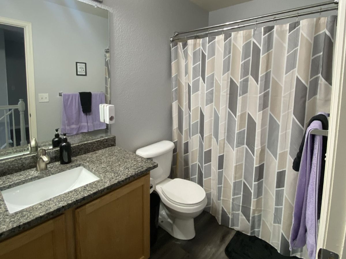 A bathroom with a granite countertop, sink, toilet, and a shower with a geometric-patterned curtain. Purple towels hang on the wall and towel rack.