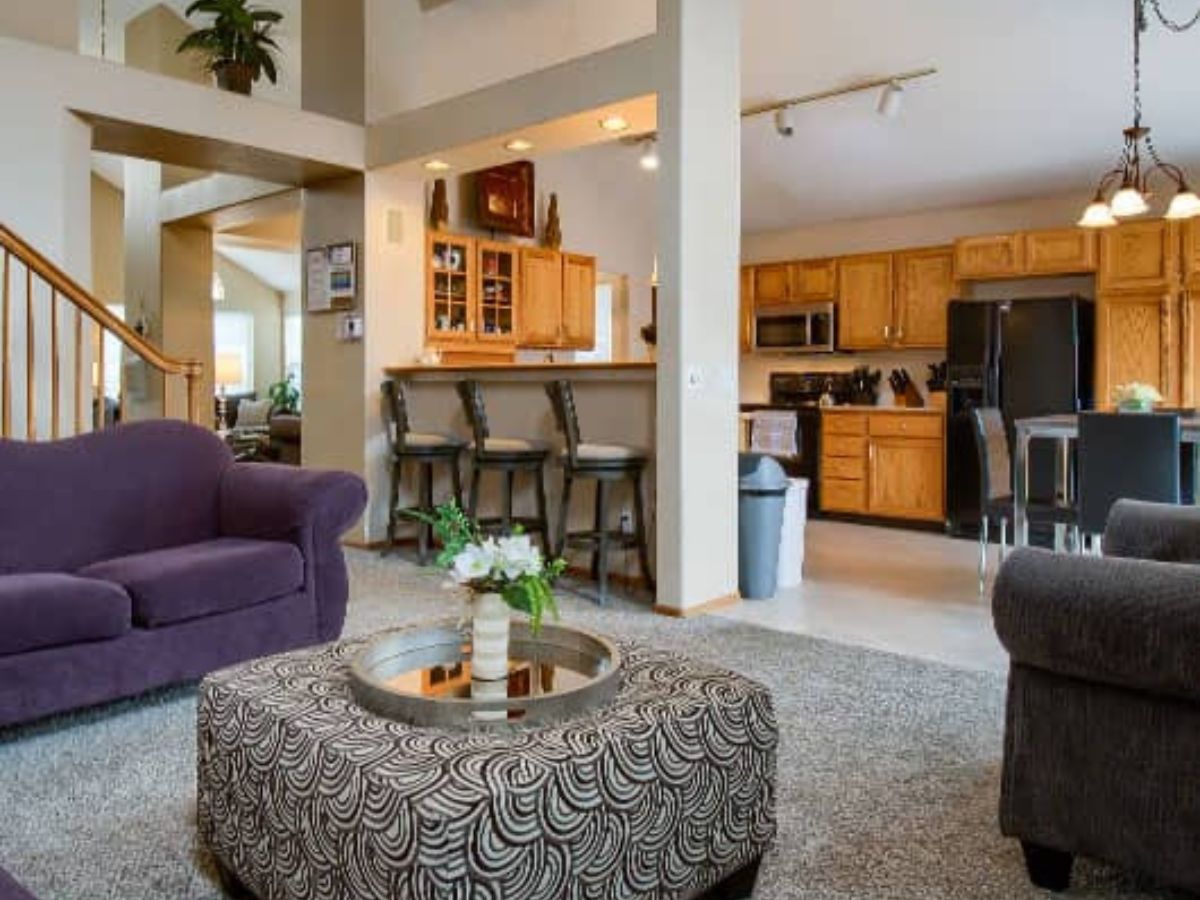 Open living room and kitchen area with purple sofas, patterned ottoman, bar stools at counter, and wood cabinetry under warm lighting.