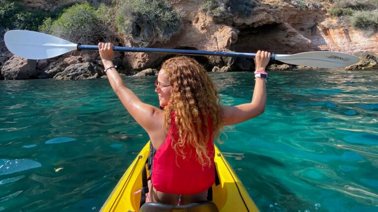 A person embracing a sober social life wears a red life vest and holds a kayak paddle above their head while sitting in a yellow kayak on clear turquoise water near rocky cliffs.