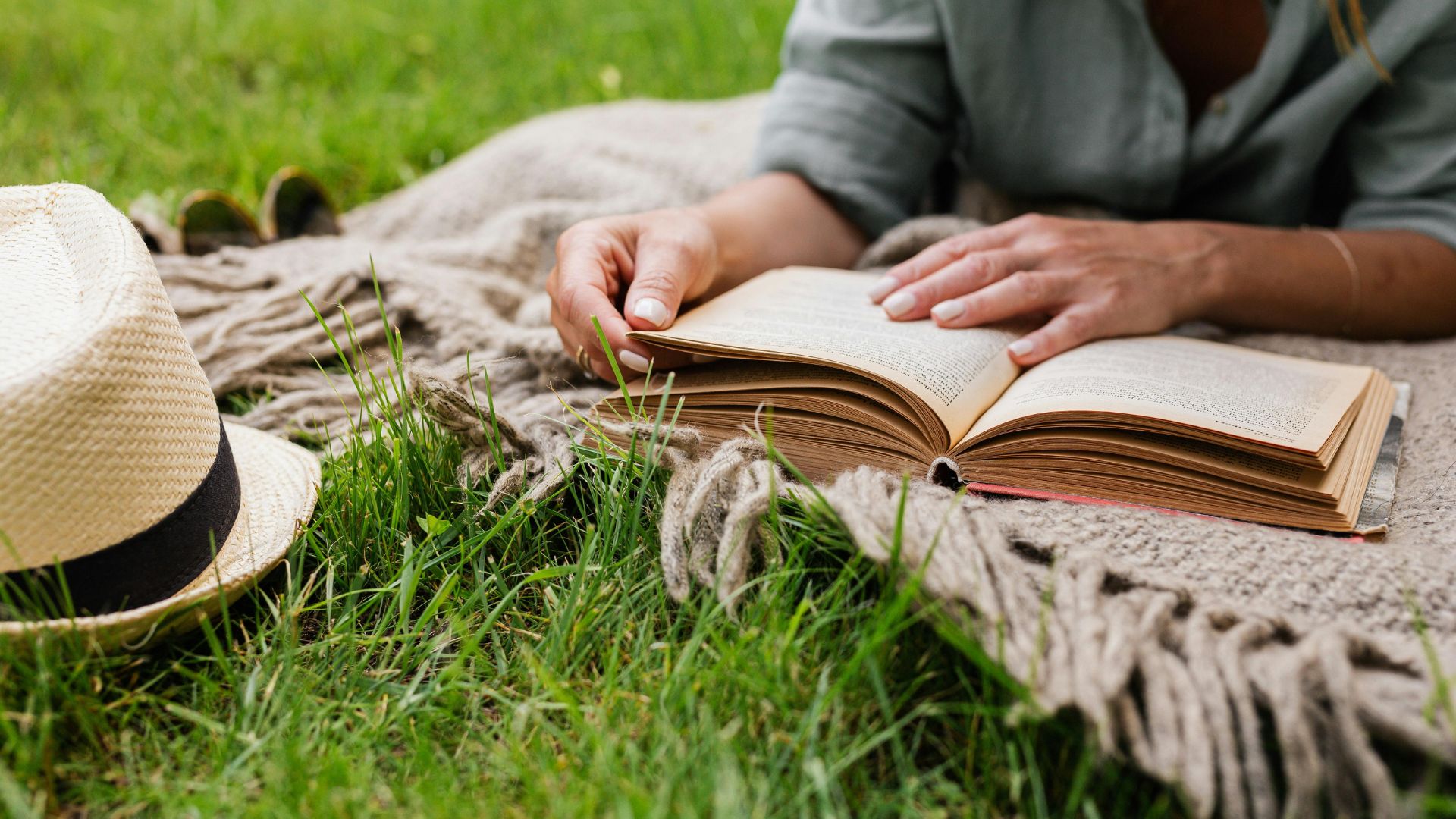 A person lies on a blanket in the grass, reading a book with a straw hat placed nearby, as they ponder life’s big questions—like what is a sober living home.