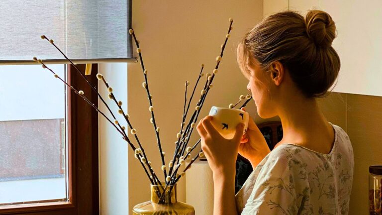 A woman with hair in a bun holds a mug and looks out a window beside a vase with pussy willow branches, contemplating financial advice for women in recovery.
