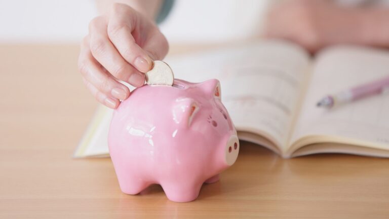 A person places a coin into a pink piggy bank on a wooden table, with an open notebook and pen in the background—perhaps jotting down sober vacation tips for their next trip.