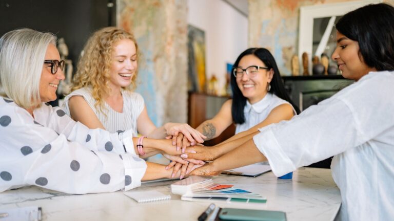 Four women sit around a table, smiling and stacking their hands together in unity, celebrating their commitment to supporting each other with financial advice for women in recovery.