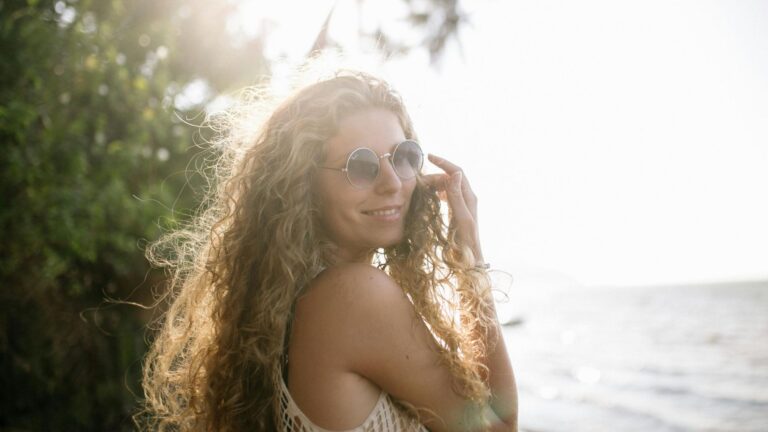 A woman with long curly hair and round sunglasses stands by the water, smiling, with sunlight shining through trees in the background—like the calm clarity found in a sober living home. Sober Summer Tips