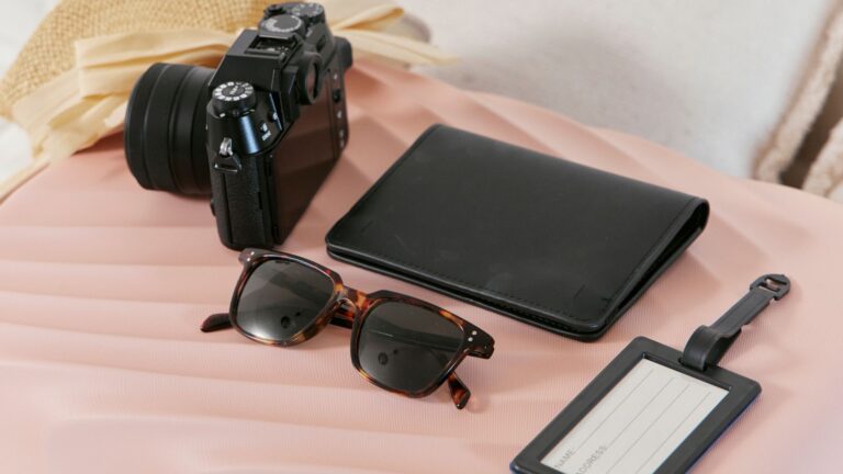 A camera, black wallet, sunglasses, and a blank luggage tag are arranged on a pink suitcase next to a straw hat—perfect inspiration for things to do instead of drinking.